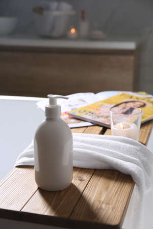 Wooden bath tray with shower gel, candle and fresh towel on tub indoors