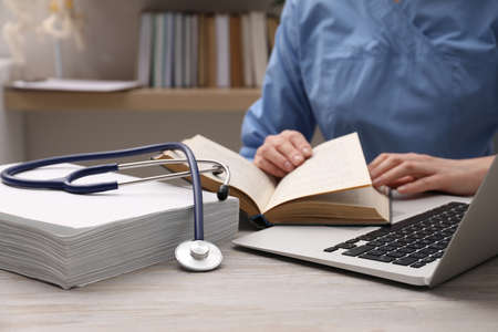 Woman with book, stethoscope and laptop at table indoors, closeup. medical education