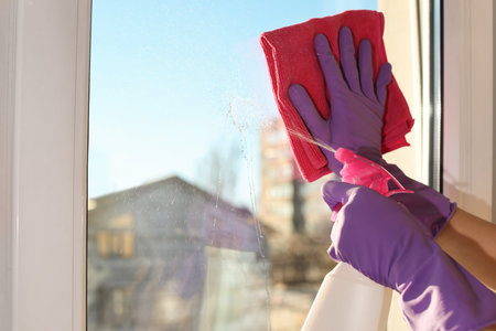 Young woman cleaning window glass with rag and detergent at home, closeup. Space for textの写真素材