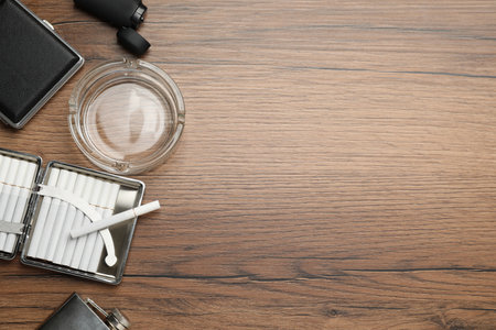 Open case with tobacco filter cigarettes, glass ashtray, lighter and pocket flask on wooden table, flat lay. Space for textの写真素材