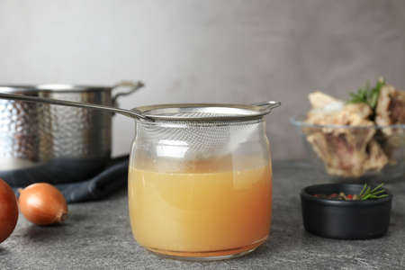 Glass jar with delicious bone broth and sieve on gray table