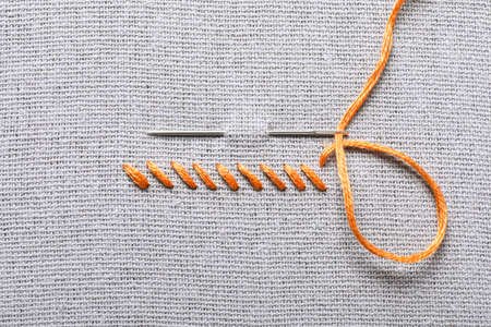 Needle with orange embroidery floss and row of stitches on gray fabric, top view