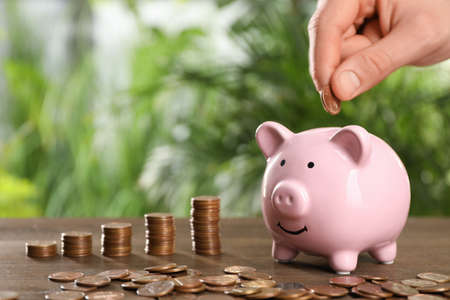 Woman coin into piggy bank at wooden table against blurred green background, closeup. Space for text