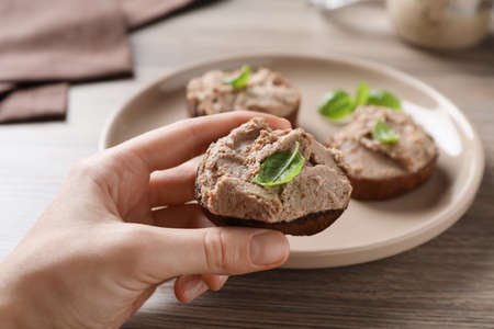 Woman taking slice of bread with delicious pate and basil from plate on wooden table, closeup
