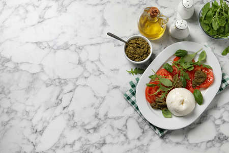 Delicious burrata salad with tomatoes, arugula and pesto sauce served on white marble table, flat...