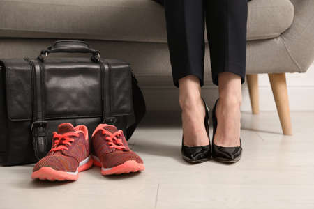 Woman in high heel shoes sitting on sofa near comfortable sneakers in office, closeupの写真素材