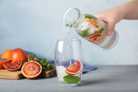 Woman pouring refreshing drink with sicilian orange from bottle into glass at gray wooden table,...