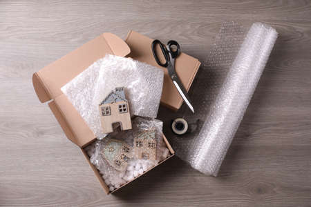 Open box with decorative house figures, adhesive tape, scissors, foam peanuts and  wrap on wooden...