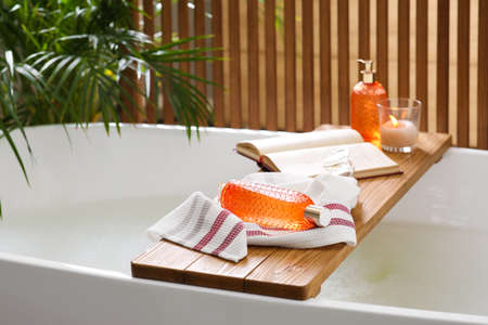 Wooden bath tray with open book, candle and body care products on tub indoors. Relaxing atmosphere