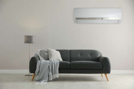 Modern air conditioner on white wall in room with stylish gray sofa