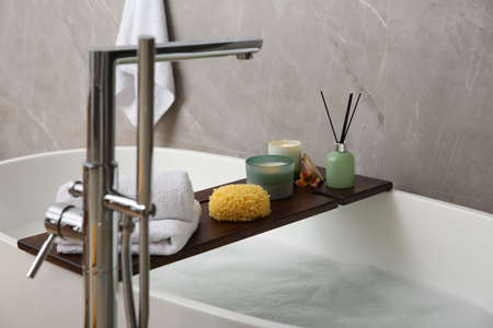 Wooden bath tray with candles, air freshener, towels and sponge on tub indoorsの写真素材