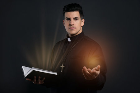 Priest in cassock holding Bible with holy light on black background