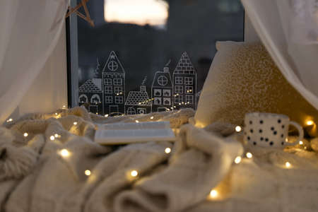 Open book and cup of hot drink on plaid near window with beautiful drawing at home. christmas decor