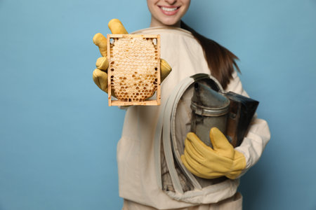 Beekeeper in uniform holding smokepot and hive frame with honeycomb on light blue background,...