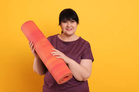 Happy overweight mature woman with yoga mat on orange background