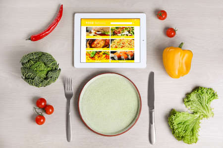 Modern tablet with open page for online food ordering, scattered vegetables, plate and cutlery on white wooden table, flat lay. Concept of delivery serviceの写真素材