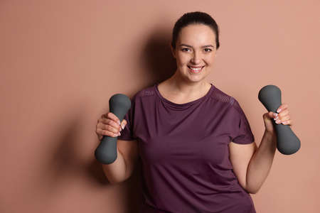 Happy overweight woman doing exercise with dumbbells on pale pink background