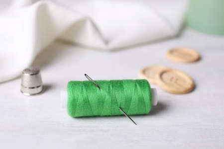 Spool of green sewing thread with needle, thimble and buttons on white table