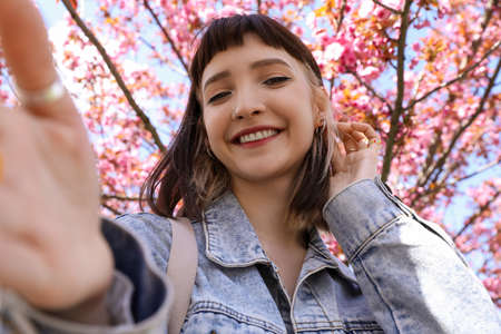 Beautiful young woman taking selfie in park with blossoming sakura treesの写真素材