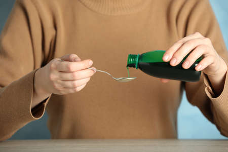 Woman pouring cough syrup into spoon at table on light blue background, closeup