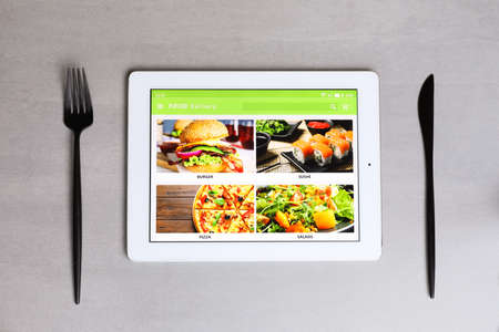 Modern tablet with open page for online food ordering and cutlery on gray table, flat lay. Concept...