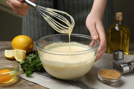 Woman making homemade mayonnaise in glass bowl at wooden table, closeup