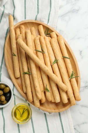 Delicious grissini, rosemary, oil and olives on white marble table, flat lay