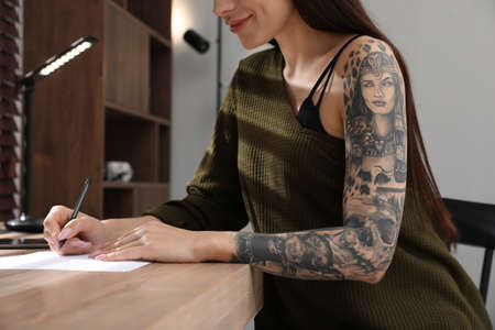Beautiful woman with tattoos on arm drawing in sketchbook at table indoors, closeupの写真素材