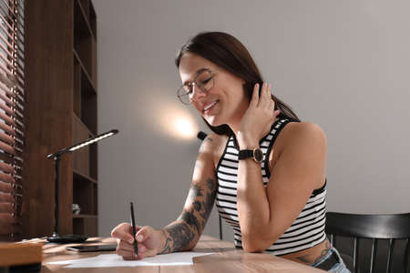 Beautiful woman with tattoos on body drawing in sketchbook at table indoors