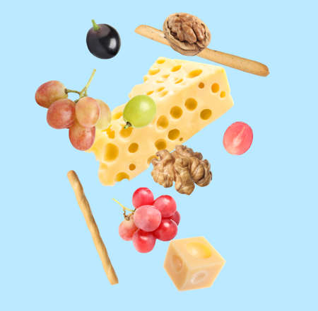 Cheese, breadsticks, grapes and walnuts falling against pale light blue background