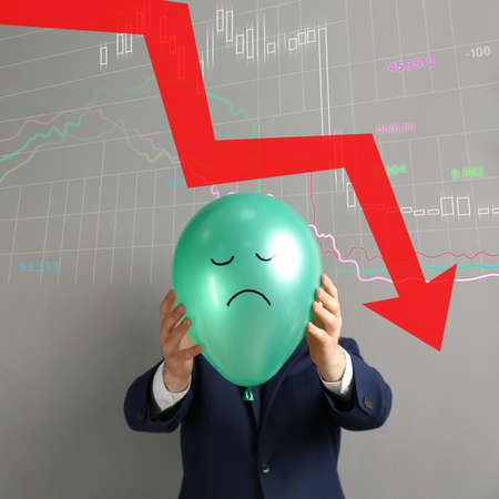 Businessman holding air balloon with drawn sad face and illustration of falling down chart on light...