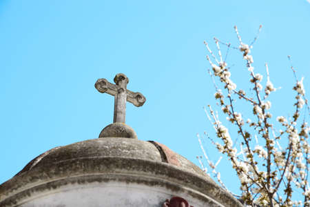 Roof of old chapel with stone cross near blossoming tree against blue sky, closeupの写真素材