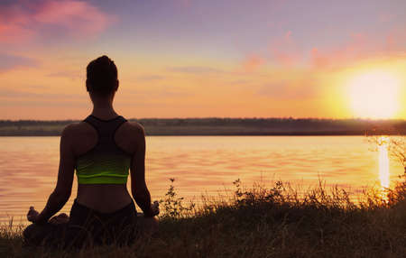 Woman meditating near river at sunset, back view. Practicing yogaの写真素材