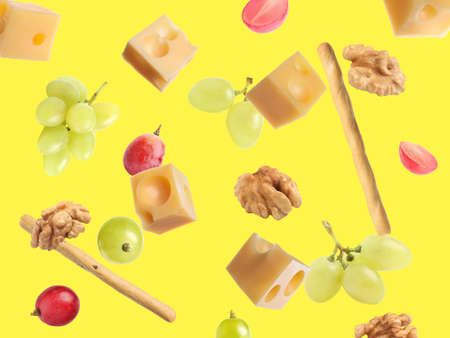 Cheese, breadsticks, grapes and walnuts falling against yellow background