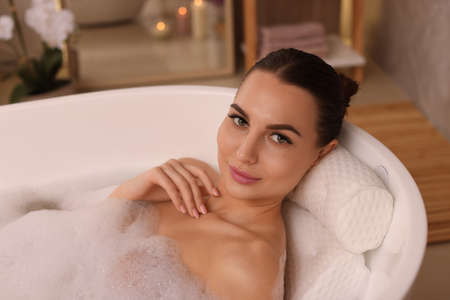 Young woman using pillow while enjoying bubble bath indoors