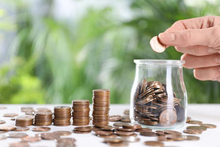 Woman coin into glass jar at white table against blurred green background, closeup. Space for textの写真素材