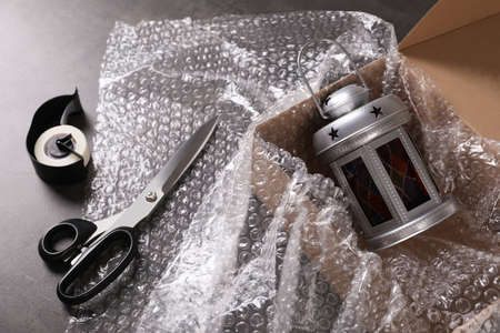 Beautiful decorative lantern, bubble wrap, scissors and cardboard box on dark gray table