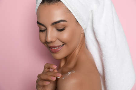 Beautiful young woman with towel applying cream on shoulder against pink backgroundの写真素材