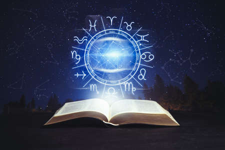 Old book on black surface and illustration of zodiac wheel with astrological signs under starry sky at nightの写真素材