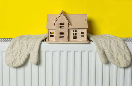 Wooden house model and knitted mittens on heating radiator near yellow wall. energy efficiency conceptの写真素材