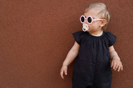 Cute little girl wearing stylish clothes with sunglasses and pacifier near brown wall. Space for textの写真素材