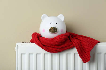 Piggy bank wrapped in scarf on heating radiator against beige backgroundの写真素材