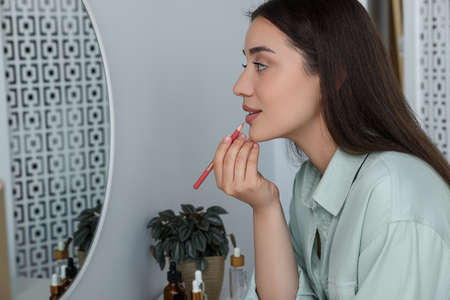 Beautiful young woman applying cosmetic pencil on lips near mirror indoorsの写真素材