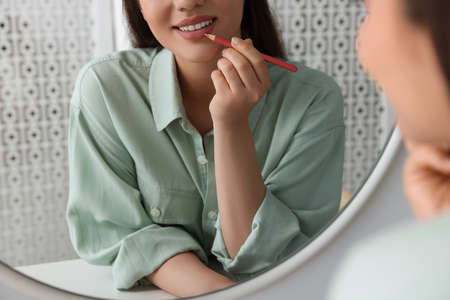 Beautiful young woman applying cosmetic pencil on lips near mirror indoors, closeupの写真素材