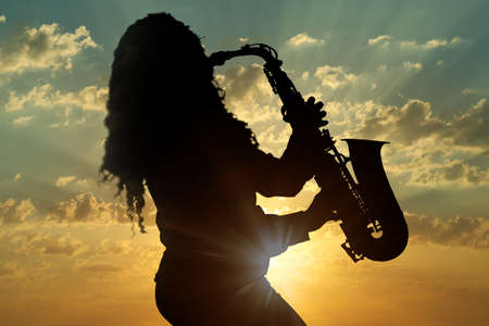 Silhouette of woman playing saxophone against beautiful sky at sunsetの写真素材