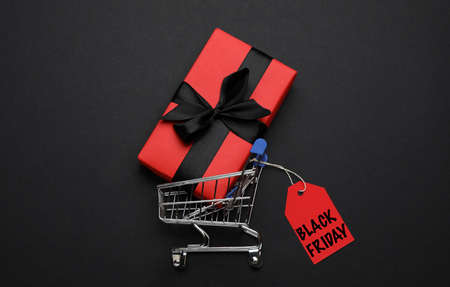 Shopping cart, tag with phrase BLACK FRIDAY and gift box on dark background, flat layの写真素材