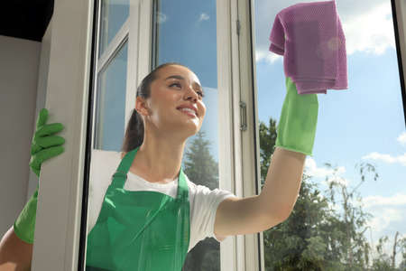 Happy young woman cleaning window glass with rag indoors