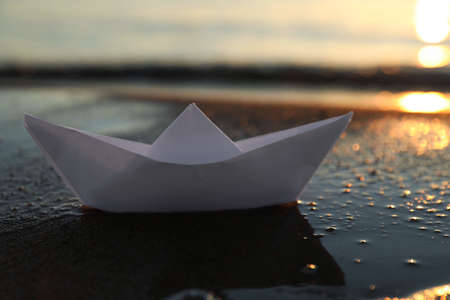 White paper boat on wet sand near sea at sunsetの写真素材