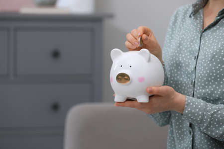 Young woman putting coin into piggy bank indoors, closeupの写真素材