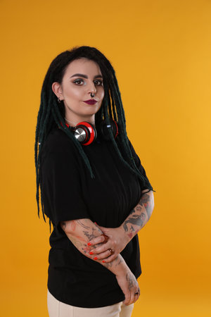 Beautiful young woman with tattoos on arms, nose piercing and dreadlocks against yellow background